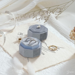 May include: Two gray velvet ring boxes, one open with a ring inside, and a gold ring with three stones. The boxes are hexagonal and sit on a cream-colored surface with decorative elements. A small pearl sits on top of the closed box.