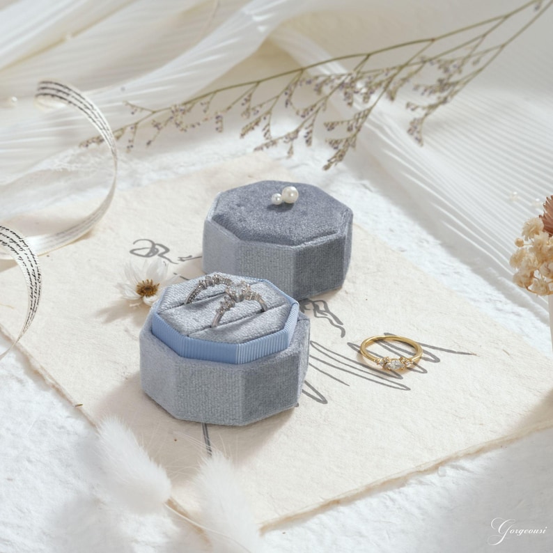 May include: Two gray velvet ring boxes, one open with a ring inside, and a gold ring with three stones. The boxes are hexagonal and sit on a cream-colored surface with decorative elements. A small pearl sits on top of the closed box.