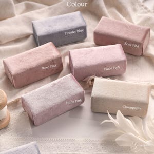 Könnte beinhalten: Eine Sammlung rechteckiger Samtboxen in verschiedenen Farben, darunter Puderblau, Rosarot, Nude Pink und Champagner. Jede Box hat ein weiches, strukturiertes Aussehen, mit den aufgedruckten Farbbezeichnungen. Die Boxen sind auf einem hellen Stoff angeordnet.