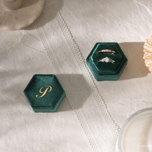 May include: Two emerald green velvet hexagon ring boxes, one with a gold initial "P" and the other with two rings. One ring is a delicate band with small stones, and the other has a cluster of stones.