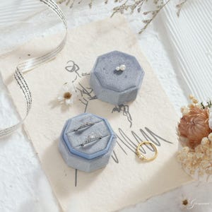 Puede incluir: Dos joyeros de terciopelo azul claro, uno abierto con anillos dentro, y un anillo de oro con peque&ntilde;as piedras. Los joyeros octagonales est&aacute;n sobre papel texturizado, con un arreglo floral y una cinta.