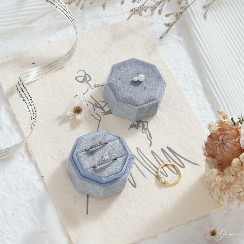 Puede incluir: Dos joyeros de terciopelo azul claro, uno abierto con anillos dentro, y un anillo de oro con peque&ntilde;as piedras. Los joyeros octagonales est&aacute;n sobre papel texturizado, con un arreglo floral y una cinta.