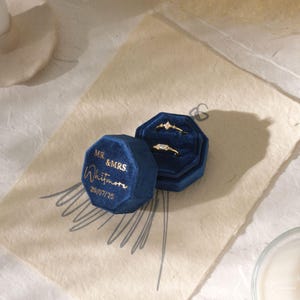 Puede incluir: Dos anillos de oro con diamantes se encuentran en un joyero de terciopelo azul oscuro. La caja est&aacute; abierta, revelando los anillos contra un interior azul. Junto a ella, una segunda caja cerrada con letras doradas que dicen "MR. & MRS. Whitmore 20/07/25".