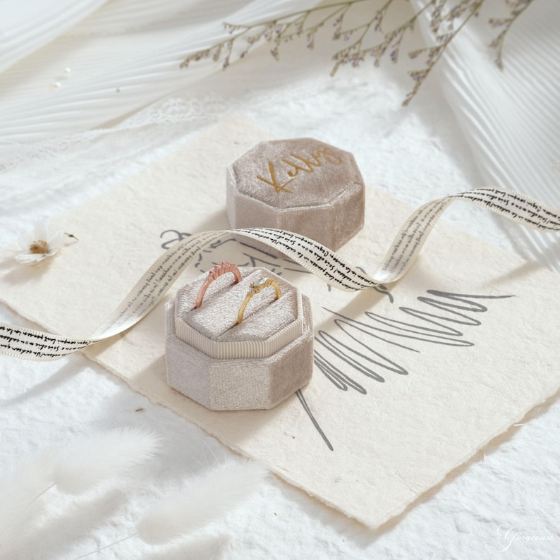 May include: Two rings, one gold and one rose gold, displayed in a light gray velvet ring box. A second ring box with the name "Kathy" in gold script is also present. The background includes a decorative ribbon and floral accents.