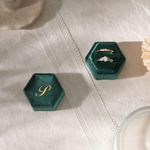 Puede incluir: Dos joyeros hexagonales de terciopelo verde esmeralda, uno con la inicial dorada "P" y el otro abierto, mostrando dos anillos. Un anillo es de oro rosa, el otro tiene un grupo de peque&ntilde;as piedras. Las cajas est&aacute;n sobre una superficie blanca con encaje.