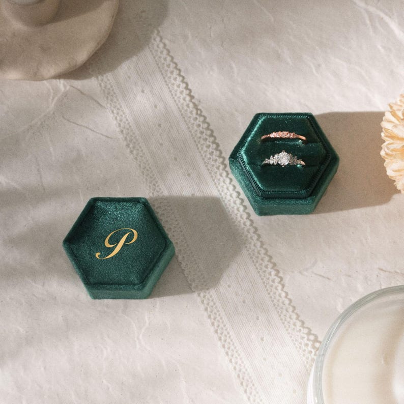 Puede incluir: Dos joyeros hexagonales de terciopelo verde esmeralda, uno con la inicial dorada "P" y el otro abierto, mostrando dos anillos. Un anillo es de oro rosa, el otro tiene un grupo de peque&ntilde;as piedras. Las cajas est&aacute;n sobre una superficie blanca con encaje.