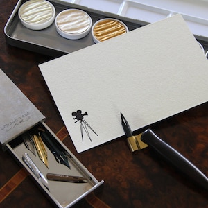 May include: A calligraphy set featuring a black oblique pen holder with a metal nib, a rectangular card with a black film camera icon, a sliding grey box labeled LONDON VALE, and a metal palette containing four circular metallic paint pans.