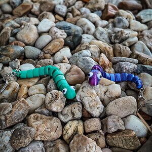 Può includere: Tre giocattoli colorati a forma di verme articolato con portachiavi poggiano su un letto di ciottoli. Uno è verde, uno è viola e blu e l'altro è blu. I giocattoli hanno grandi occhi da cartone animato e sono di plastica.