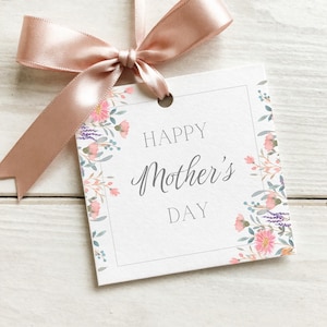 May include: A white gift tag with the words "HAPPY Mother's DAY" in elegant script. The tag is adorned with floral illustrations in pink, purple, and green, and is tied with a blush pink satin ribbon.