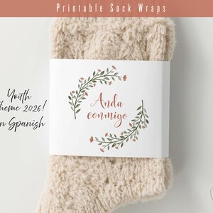 May include: A pair of beige socks with a white label featuring floral accents and the Spanish phrase "Anda conmigo." The label also includes the text "Youth theme 2026! in Spanish."