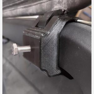 Puede incluir: Primer plano de un soporte de baca de coche de plástico negro con un tornillo plateado. El soporte está diseñado para fijarse a la baca de un coche.