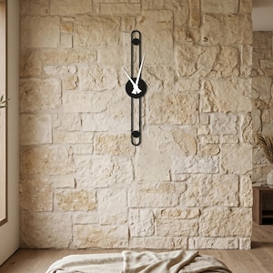 Puede incluir: Un reloj de pared negro moderno con manecillas blancas, montado en una pared de piedra texturizada. El reloj tiene una esfera circular y un diseño vertical y alargado. La habitación presenta una paleta de colores neutros.