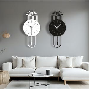Könnte beinhalten: Zwei moderne Wanduhren, eine weiße und eine schwarze, werden an einer grauen Wand gezeigt. Jede Uhr hat ein kreisförmiges Zifferblatt mit Zeigern und ein Pendeldesign. Die Uhren befinden sich über einem weißen Ecksofa.