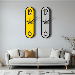 Könnte beinhalten: Zwei moderne Wanduhren mit schwarzen Zeigern und Ziffern. Eine Uhr ist gelb, die andere grau, beide mit schwarzen Rändern. Die Uhren sind über einem weißen Sofa mit Kissen montiert. Ein kleiner Beistelltisch aus Holz und eine Vase sind ebenfalls zu sehen.