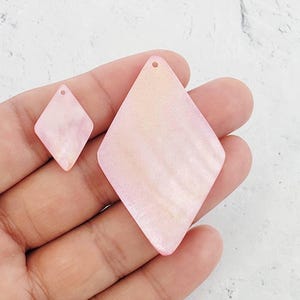 May include: Two pink iridescent diamond-shaped shell pendants with holes for hanging. The larger pendant is about 2 inches in diameter and the smaller pendant is about 1 inch in diameter.