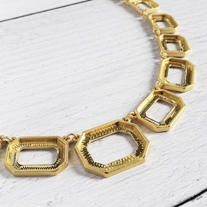 May include: A gold-toned necklace with geometric openwork rectangular and octagonal shapes. The necklace is on a white wooden surface. The chain is delicate, and the pendant shapes are connected by small links.