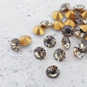 May include: A collection of small, round, faceted rhinestones in a dark gray color. The rhinestones are scattered on a white surface. Some of the rhinestones have a gold backing.