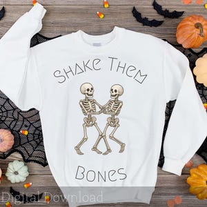 May include: White sweatshirt with the text "Shake Them Bones" above a graphic of two dancing skeletons. The design is in black and tan. The image is surrounded by Halloween decorations, including pumpkins and candy corn.