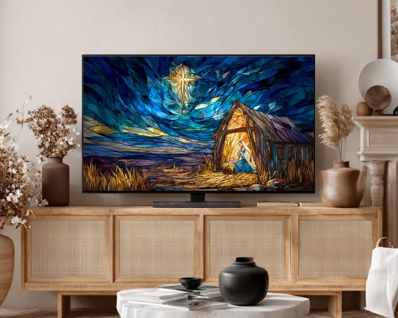 May include: A flat-screen television displays a stained-glass style image of a nativity scene. The artwork features a star, a stable, and figures in blue and gold against a dark blue sky. The TV sits on a light wood cabinet.