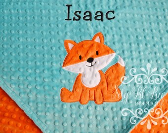 May include: A baby blanket with a teal blue minky dot fabric and an orange minky dot fabric on the reverse side. The blanket features an embroidered orange fox with white accents and the name "Isaac" in black thread.