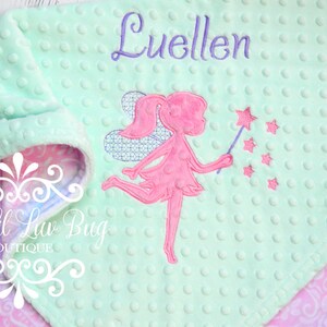 May include: A mint green baby blanket with a pink fairy design and the name "Luellen" embroidered in purple. The blanket has a textured surface and a patterned lining in pink and purple.