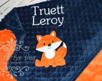 May include: A navy blue dimpled minky blanket featuring an embroidered orange fox with a white belly and tail tip. The name Truett Leroy is embroidered in white thread above the fox. The blanket has a bright orange underside and a soft texture.