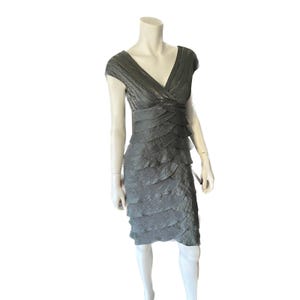 Puede incluir: Un vestido gris, hasta la rodilla, con cuello en V y mangas cortas. El vestido presenta una falda con volantes en capas y una cintura ceñida. La tela tiene una apariencia texturizada, y el vestido se exhibe en un maniquí.