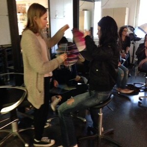 May include: Two people are sitting on stools in a room. The person on the left is wearing a light brown sweater and the person on the right is wearing a black leather jacket and blue jeans. The person on the right is holding a pink and white object.