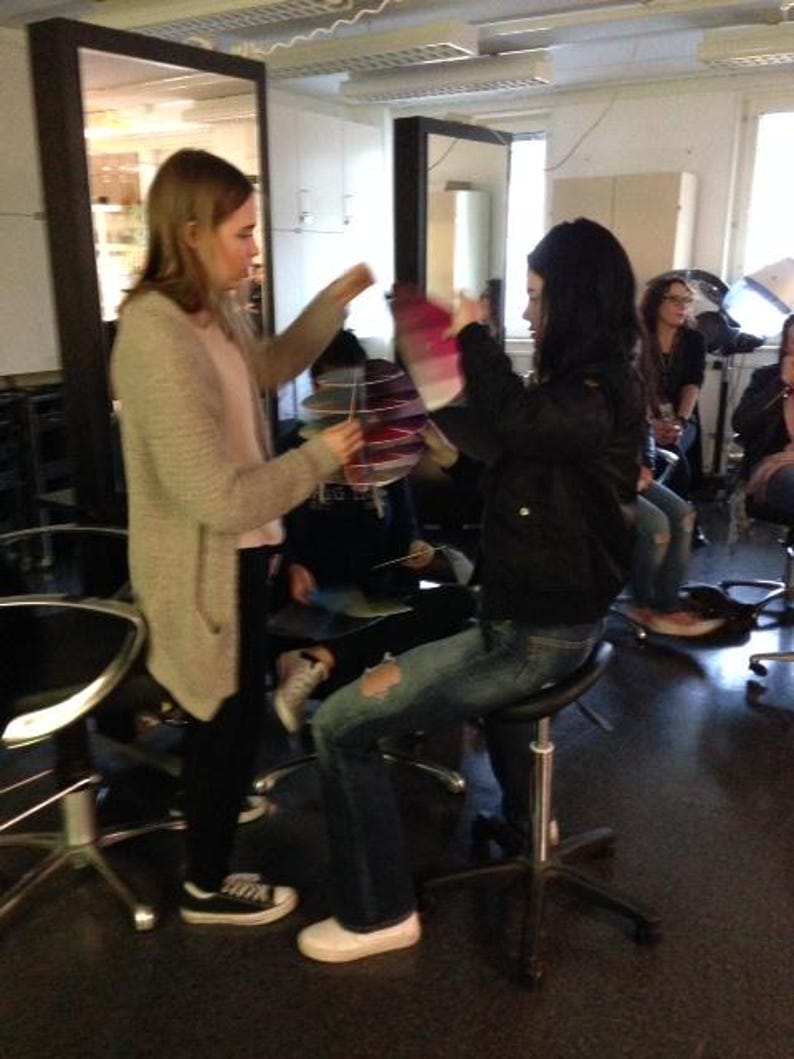 May include: Two people are sitting on stools in a room. The person on the left is wearing a light brown sweater and the person on the right is wearing a black leather jacket and blue jeans. The person on the right is holding a pink and white object.