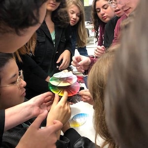 May include: A group of people are gathered around a table, looking at a color wheel. The color wheel is made up of several circular pieces of paper, each with a different color. The people are all wearing casual clothing and appear to be students.