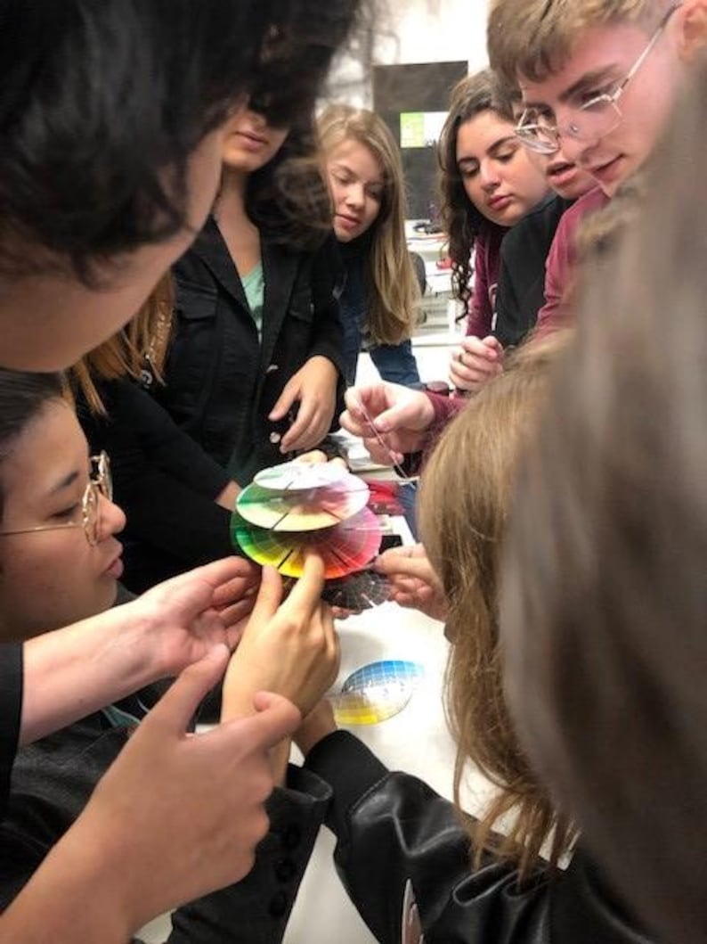 May include: A group of people are gathered around a table, looking at a color wheel. The color wheel is made up of several circular pieces of paper, each with a different color. The people are all wearing casual clothing and appear to be students.