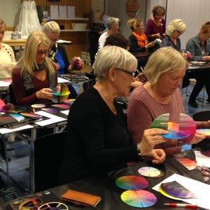 May include: A group of people are gathered around tables, each holding a color wheel. They are looking at the color wheels and discussing them. The color wheels are a variety of colors, including red, orange, yellow, green, blue, indigo, and violet.