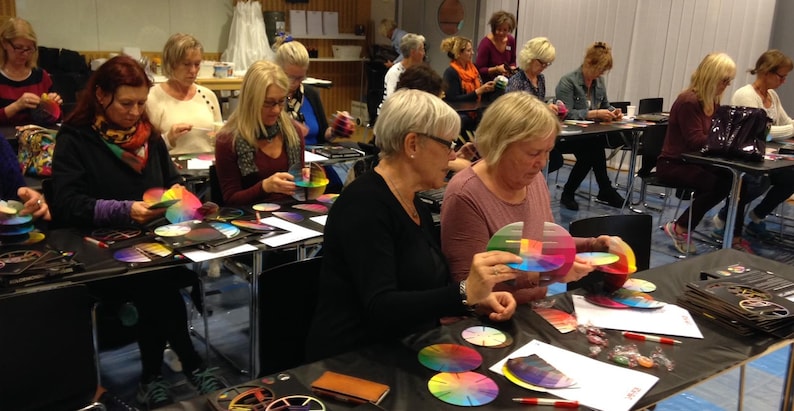 May include: A group of people are gathered around tables, each holding a color wheel. They are looking at the color wheels and discussing them. The color wheels are a variety of colors, including red, orange, yellow, green, blue, indigo, and violet.