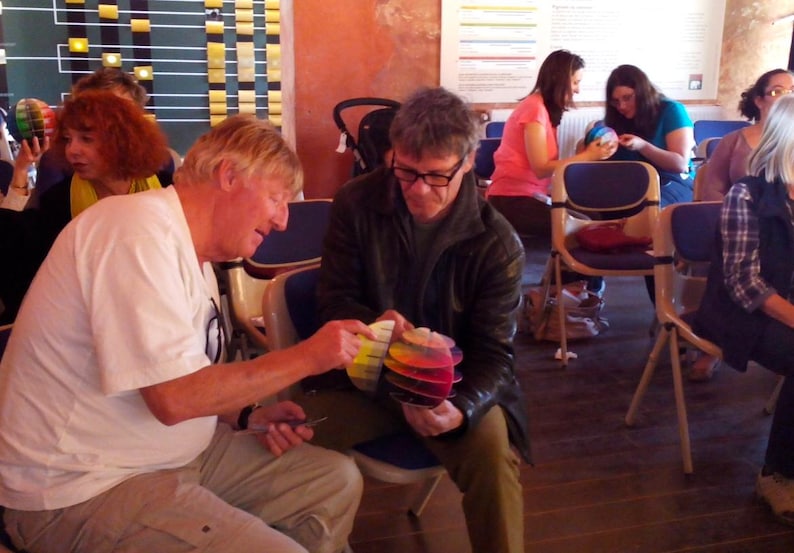 May include: A group of people sitting in chairs, with one person holding a colorful stack of paper. The paper is arranged in a circular pattern, with each layer a different color.