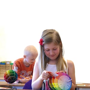 May include: Two children are sitting at a table. One child is using a pencil to write on a piece of paper. The other child is holding a color wheel and looking at it. The color wheel is made of several colorful circles.