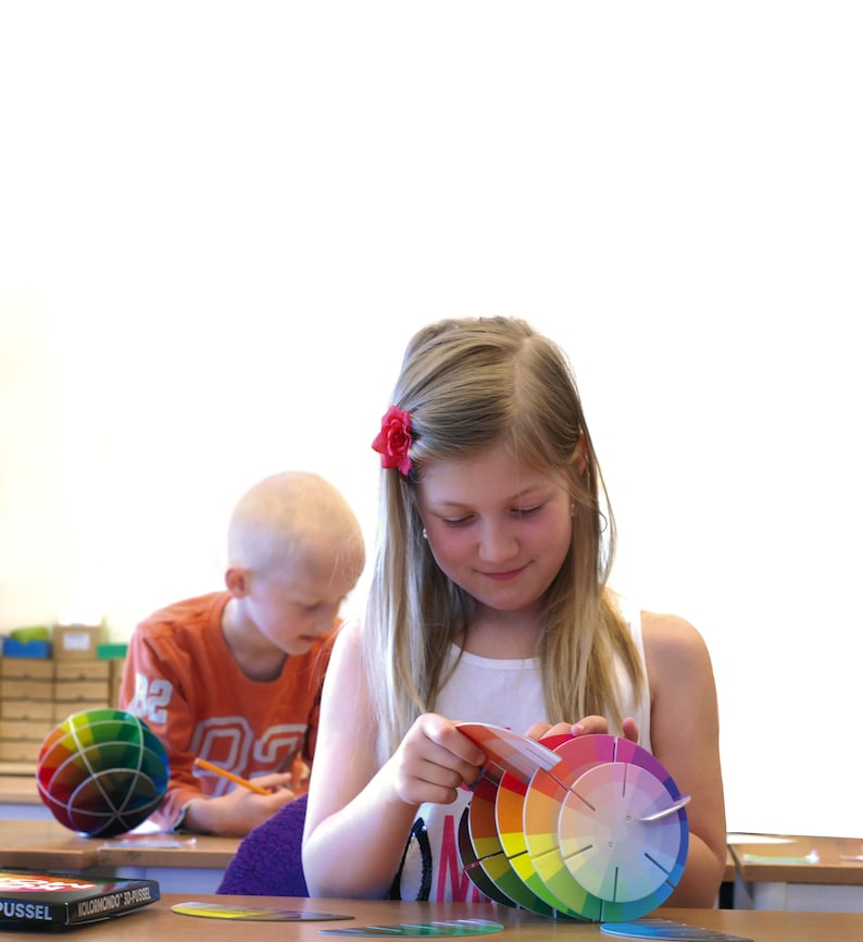 May include: Two children are sitting at a table. One child is using a pencil to write on a piece of paper. The other child is holding a color wheel and looking at it. The color wheel is made of several colorful circles.
