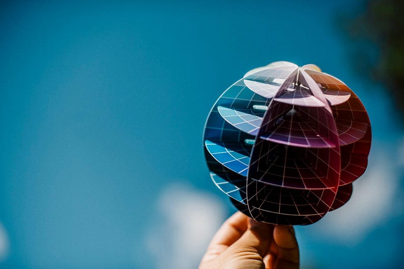 May include: A colorful, geometric globe made of paper, with a black grid pattern and a gradient of blue, purple, and red colors. The globe is held in a hand against a blue background.