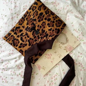 Könnte beinhalten: Eine Clutch mit Leopardenmuster, einem dunkelbraunen Riemen und einer kleinen schwarzen Schleife. Eine florale Karte ist im Inneren zu sehen. Die Clutch liegt auf einer weißen Oberfläche mit Blumenmuster.