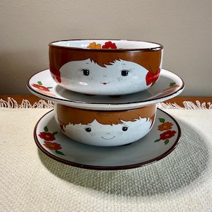 May include: Two ceramic bowls and saucers stacked. The bowls feature a child's face with brown hair and a smiling expression. The saucers and the rim of the bowls have a brown border. Floral designs in red and orange decorate the saucers and the inside of the bowls.