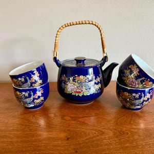 May include: A cobalt blue ceramic tea set consisting of a teapot with a woven rattan handle and four matching tea cups. Each piece features a detailed, colorful floral pattern and a peacock motif with gold accents painted on the exterior surface.