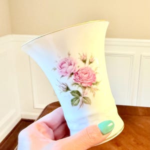 May include: Cream-colored porcelain vase with a gold rim and a flared top. The vase features a hand-painted design of pink roses with green leaves. The vase is held in a hand.