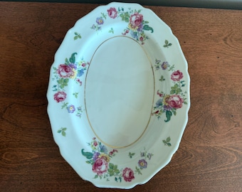 May include: An oval, off-white serving platter with a scalloped edge. The platter is decorated with a floral pattern featuring pink roses, blue and yellow flowers, and green leaves. A gold trim outlines the inner oval.