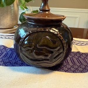 May include: A handcrafted ceramic jar with a lid. The jar is a dark brown color with a glossy finish and a decorative embossed design around the middle. The lid and the top of the jar are a lighter brown color.