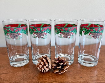 May include: A set of four clear glass tumblers, each featuring a decorative band near the rim with a red background, green ribbon, holly leaves, and brown pinecones. Two natural brown pinecones are positioned in front of the glasses.