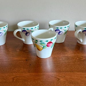 May include: Set of eight white ceramic mugs with a decorative fruit and leaf design. Each mug features a handle and is adorned with painted plums, pears, and tulips in shades of purple, yellow, red, and green.