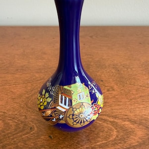 May include: A small, cobalt blue vase with a gold rim. The vase features a detailed, hand-painted scene of a carriage with yellow, green, and brown accents. The vase is approximately 5 inches tall.