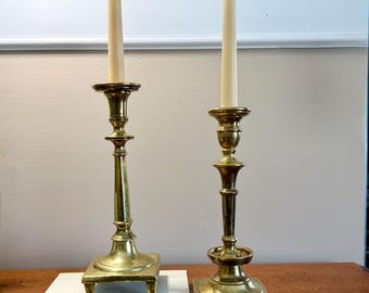 May include: Two antique brass candlesticks with tall, slender ivory candles. The candlesticks have ornate bases and detailed stems. A small bouquet of white flowers and greenery rests on a wooden surface in front of the candlesticks.