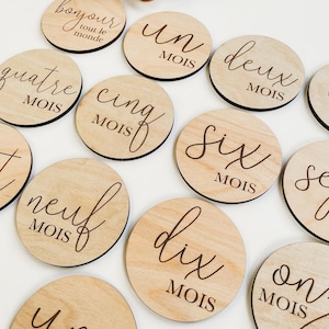 May include: Wooden baby milestone discs with French text, including "bonjour tout le monde", "un mois", "deux mois", "quatre mois", "cinq mois", "six mois", "neuf mois", "dix mois", "onze mois", and "une an".