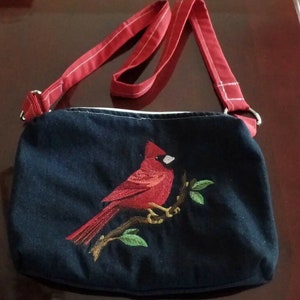 May include: A black canvas crossbody bag with a red cardinal embroidered on the front. The bag has a red adjustable strap.
