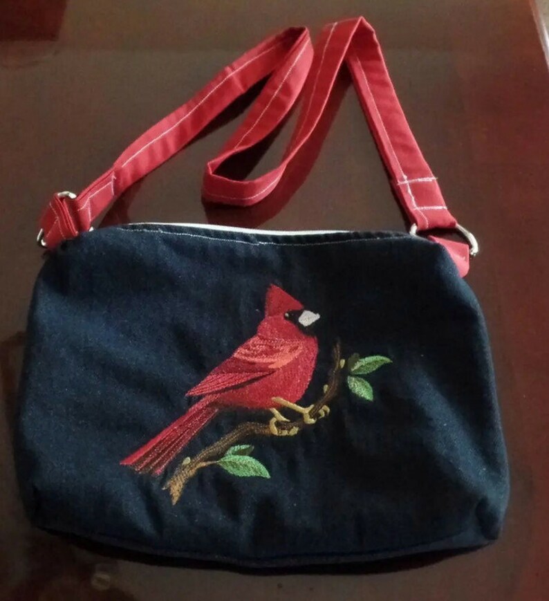 May include: A black canvas crossbody bag with a red cardinal embroidered on the front. The bag has a red adjustable strap.
